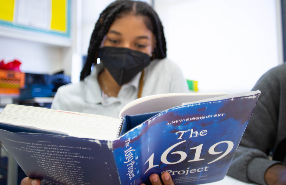 A high school student wearing a mask reads from the book the 1619 project